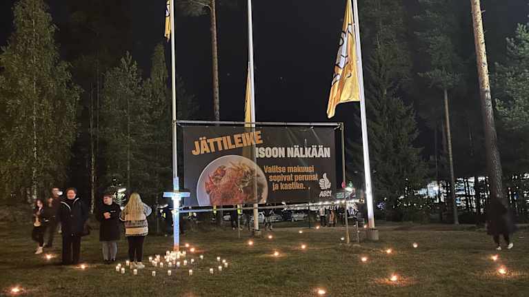 Pihamaalla iso mainoskyltti, jonka ympärillä liput ovat puolitangossa ja nurmikolla palaa ulkotulia. 