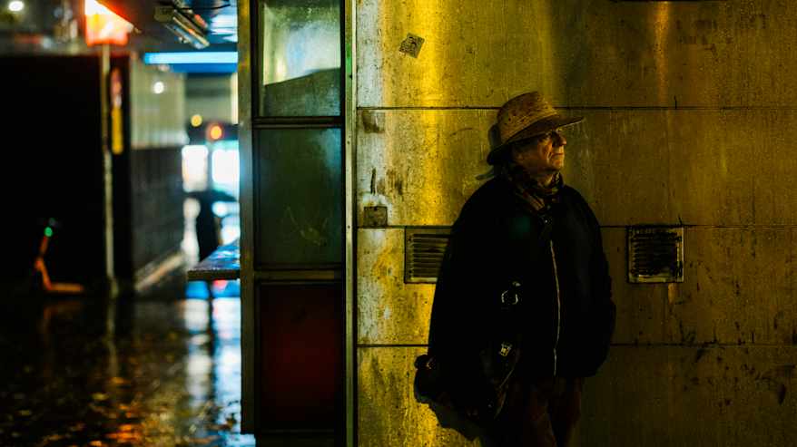 Henkilö seisoo iltahämärässä kadulla, jonka valaistus heijastuu märästä pinnasta. 