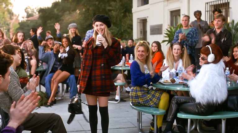 Kuvassa näkyy Clueless-elokuvan päähenkilö, joka on pukeutunut tyylikkääseen asuun. Hän on koulun pihalla, oppilaat taputtavat, hän on keskipiste.