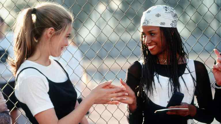 Clueless-elokuvan kaksi päähenkilöä tervehtivät hymyillen urheilukentällä, taustalla verkkoaita.