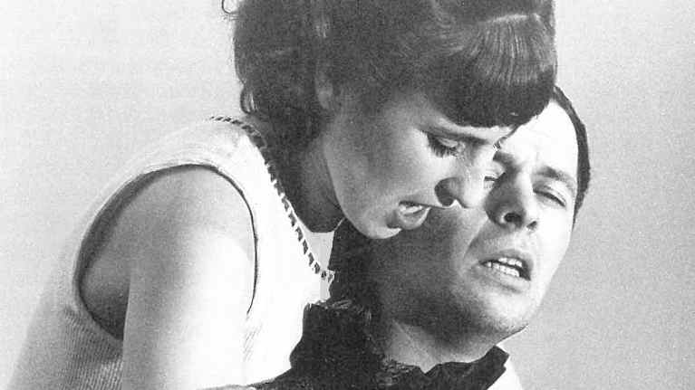 Kaksi henkilöä poseeraa yhdessä muodollisissa ja tyylikkäissä vaatteissa nostalgisessa mustavalkovalokuvassa.