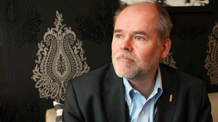 Mies istuu hotellin aulassa, pukeutuneena pukuun ja kauluspaitaan,