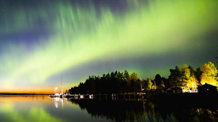 Revontulia taivaalla. Keskellä ja oikealla alhaalla näkyy laituri, rakennuksia ja valoja. 