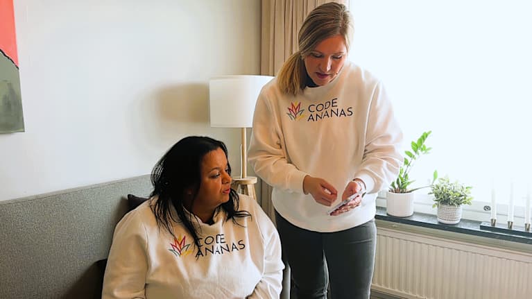 Kaksi naista keskustelevat olohuoneessa, toinen heistä selaa puhelintaan.