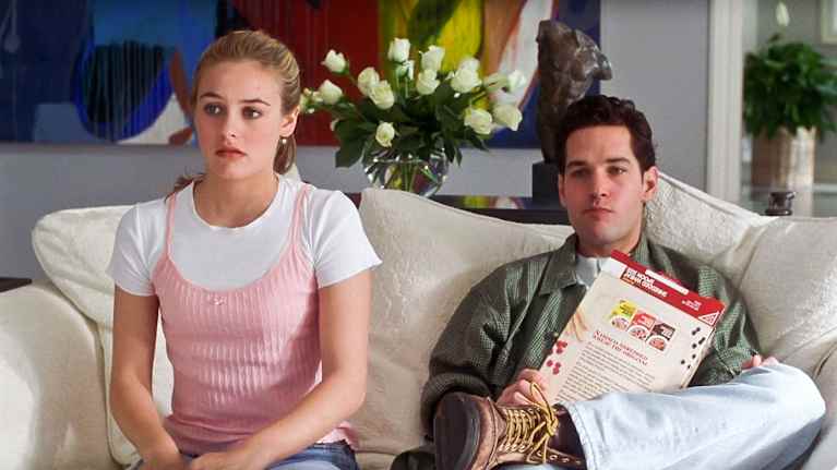 Clueless-elokuvan kaksi päähenkilöä, nuori mies ja nainen, istuvat vaalealla sohvalla, katsovat televisiota. Taustalla on värikäs maalaus ja kukkia maljakossa.