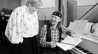 Mies istuu pianon ääressä ja pitää käsissään nautteja. Hän katsoo takanaan seisovaa naista ja hymyilee. Nainen nauraa.