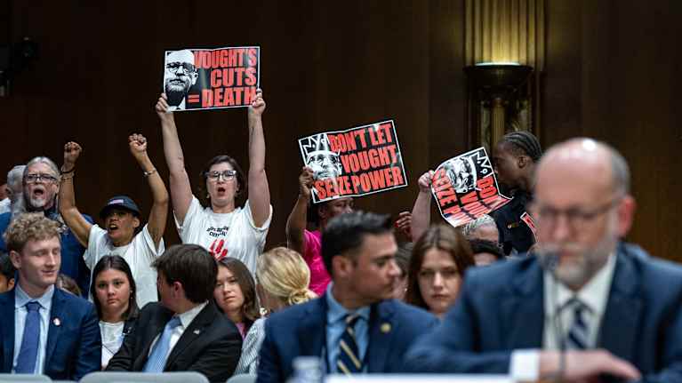Mielenosoittajat heiluttavat Voughtia vastustavia kylttejä, Vought etualalla, senaatin kuulemistilaisuus.
