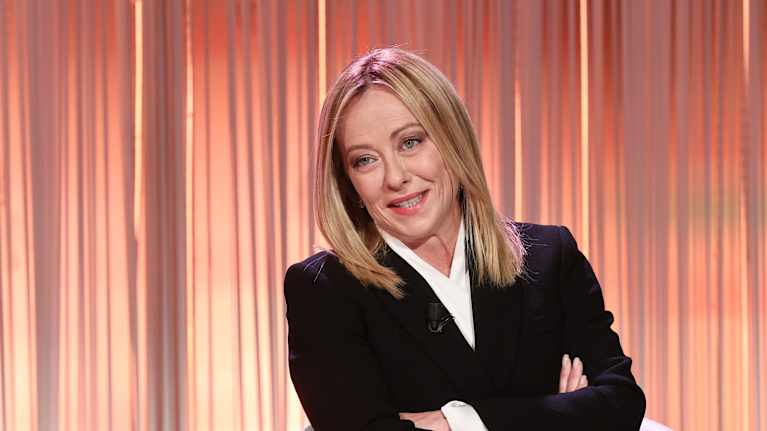 Vaalea jakkupukuinen nainen hymyilee studiovalaistuksessa kädet puuskassa vasempaan suuntaan kuvassa.