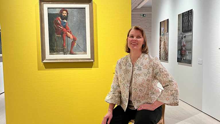 Nainen istuu jakkaralla ja hänen takanaan on keltaisella seinällä taulu. Taulussa on punavaatteinen mies.