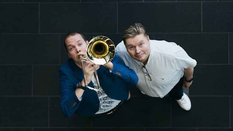 Kaksi miestä, joista toisella huulillaan pasuuna, kuvattuna ylhäältä päin.