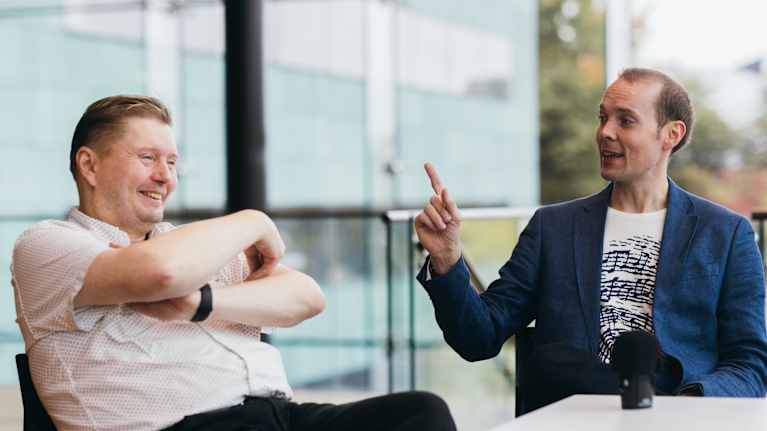 Kaksi miestä istuu pöydän ääressä, toinen hymyilee kädet puuskassa ja toinen puhuu etusormi pystyssä.