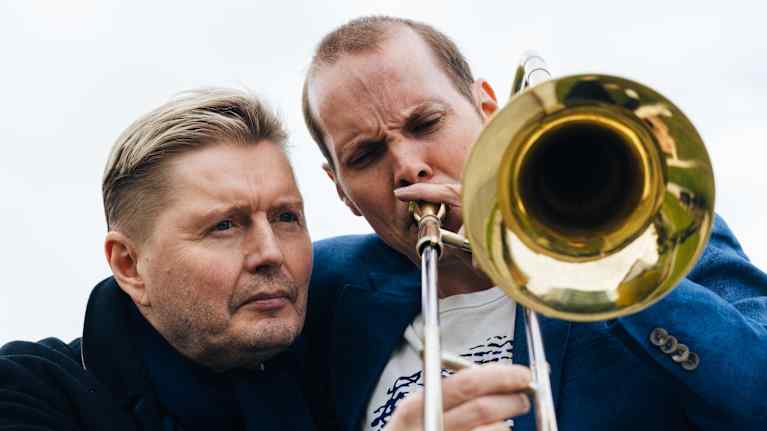 Lähikuva kahdesta miehestä, joista toinen puhaltaa pasuunaan ja toinen pitää kiinni sen luistista.
