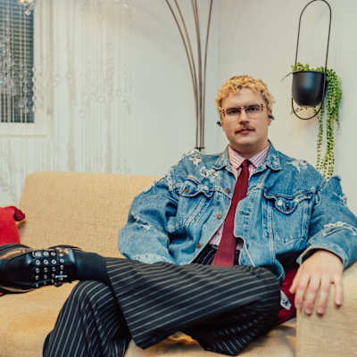 Henkilö istuu sohvalla rennosti pukeutuneena tyylikkääseen asuun, jossa yhdistyy farkkutakki, punainen kauluspaita ja raidalliset housut. Vierellä on viherkasvi, joka tuo luonnonläheistä tunnelmaa huoneeseen.