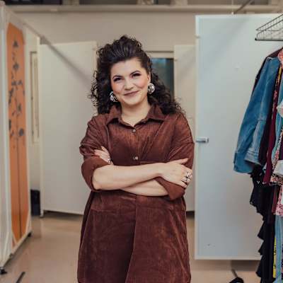 Suvi Teräsniska, pukeutuneena ruskeaan mekkoon. Hänellä on kädet ristissä rinnalla, ja taustalla näkyy hengarissa roikkuvia vaatteita sekä osia huoneesta, jossa hän on.