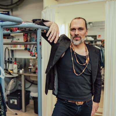 JP Leppäluoto eisoo vaaterekistä nojaten vaateliikkeessä tai vaatehuoneessa. Hänellä on yllään tumma paita, jakku ja useita koruja. Taustalla näkyy erilaisia vaatteita ja laukkuja.