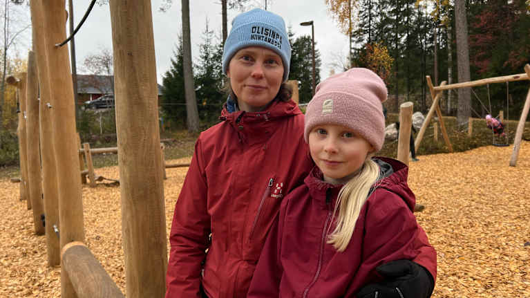 Nainen ja tyttö seisovat vierekkäin syksyisessä leikkipuistossa.