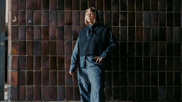 Kuvassa henkilö poseeraa seinän edessä pukeutuneena tummaan ja rentoon asukokonaisuuteen.