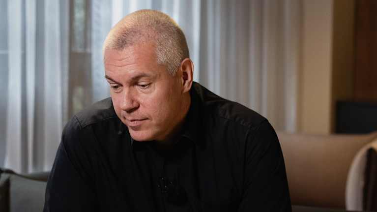 Kuvassa on mies pukeutuneena mustaan asuun, istuen sohvalla kädet ristissä edessään. Taustalla näkyvät suuret ikkunat, joista tulvii luonnonvaloa huoneeseen.