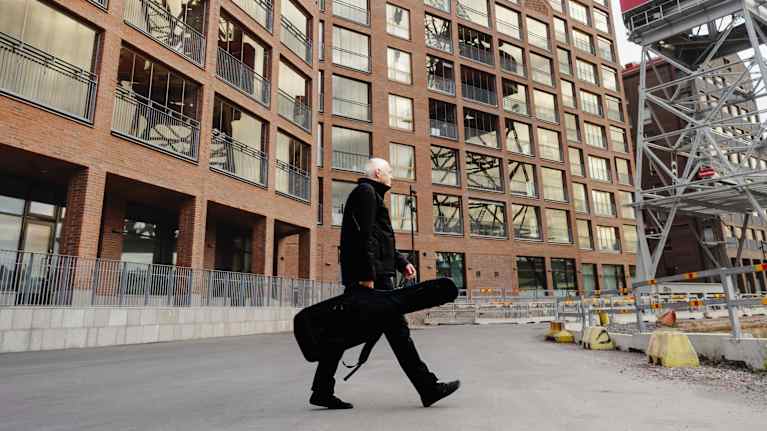 Mies kävelee arvokkaan rakennuskompleksin edustalla kantamassa soitinkoteloa. Oikealla näkyy rakennuksen osana oleva suuri nosturi, joka luo kontrastia moderniin arkkitehtuuriin.