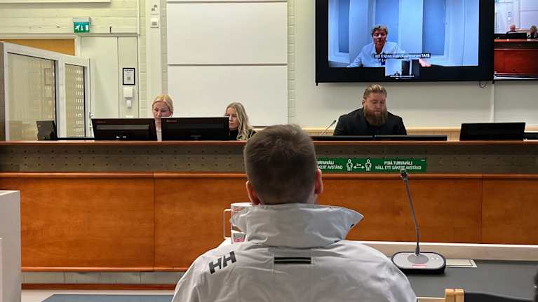 Lyhythiuksinen nuori mies istuu Kainuun käräjäoikeuden istunnossa valkoinen tuulitakki yllään.