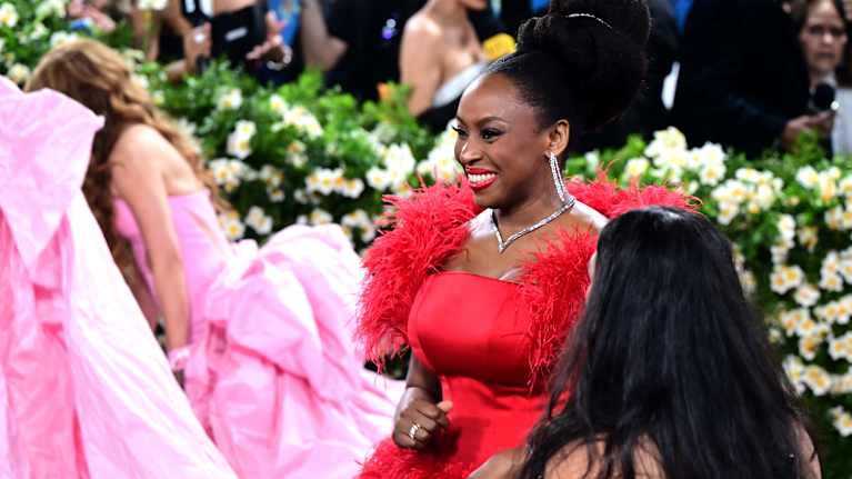 Punaiseen iltapukuun pukeutunut nainen. Puvussa on näyttäviä sulkaisia yksityiskohtia. Taustalla näkyy kukkia.