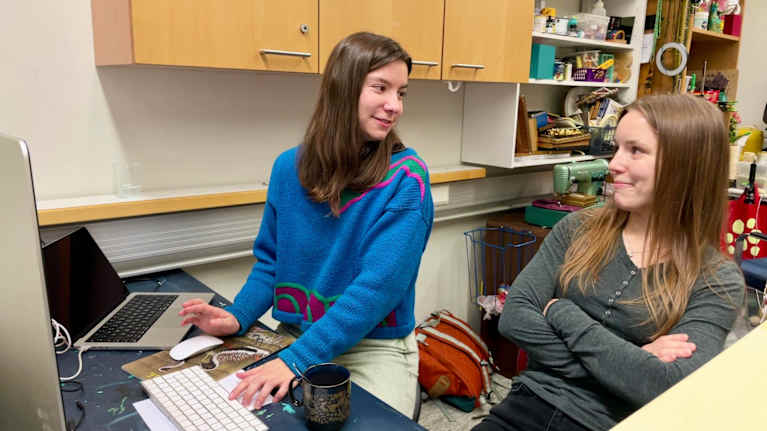 Kaksi nuorta naista istuu tietokoneen ääressä ja katsoo toisiaan hymyillen.