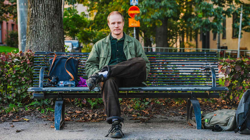 Leo Minkkinen istuu puiston penkillä ja pitää jalkaansa toisen päällä. Vieressään hänellä on reppu, kuulokkeet ja vesipullo.