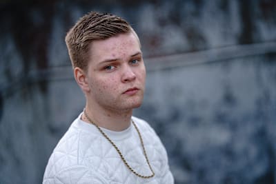 Vakavan näköinen vaalea nuorimies katsoo kameraan. Kaulassa roikkuu kultainen ketju.