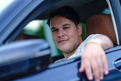 Nuori tummahiuksinen mies istuu autossa ja katsoo kameraan hymyillen. Käsi nojaa auton ikkunalla.