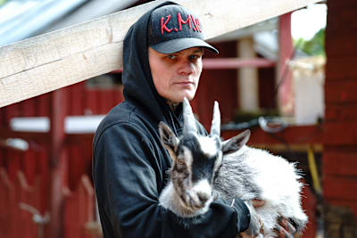 Nuori, huppupäinen mies katsoo vakavana kameraan. Miehen sylissä on vuohi. Sormet ovat täynnä tatuointeja.