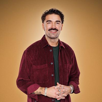 Dani Hänninen on pukeutunut viininpunaiseen samettipaitaan ja tummiin housuihin. Hänellä on useita rannekoruja ja sormus, ja hän pitää käsiään yhdessä vartalon edessä. Tausta on yhtenäisen ruskea.