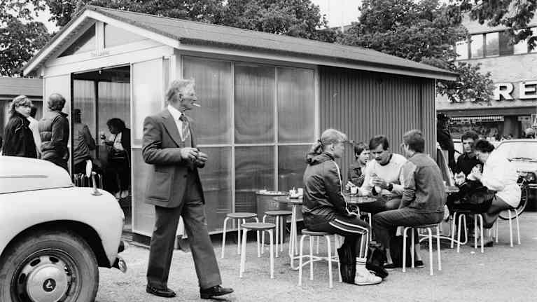 Savuketta polttava pukuun pukeutunut mies kävelee vetykioskin edessä vuonna 1983.