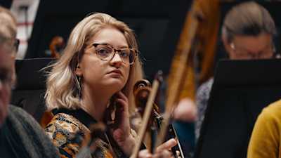 Silmälasipäinen nuori viulisti keskittyy kuuntelemaan kapellimestaria.