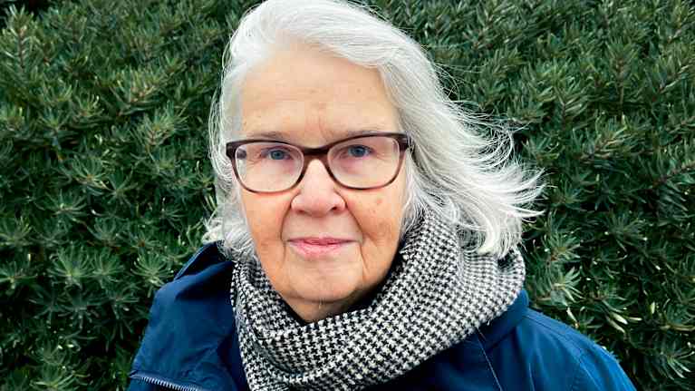 Henkilö seisoo ulkona vihreän pensaan edessä.