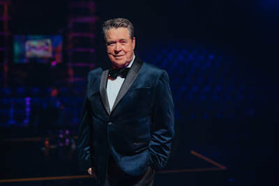 Mies pukeutunut tummansiniseen smokkiin seisoo hämärästi valaistussa tilassa, joka näyttää olevan teatteri tai esitysareena. Taustalla näkyy katsojia ja lavarakenteita. Markku Aro ja Elämäni Biisi.