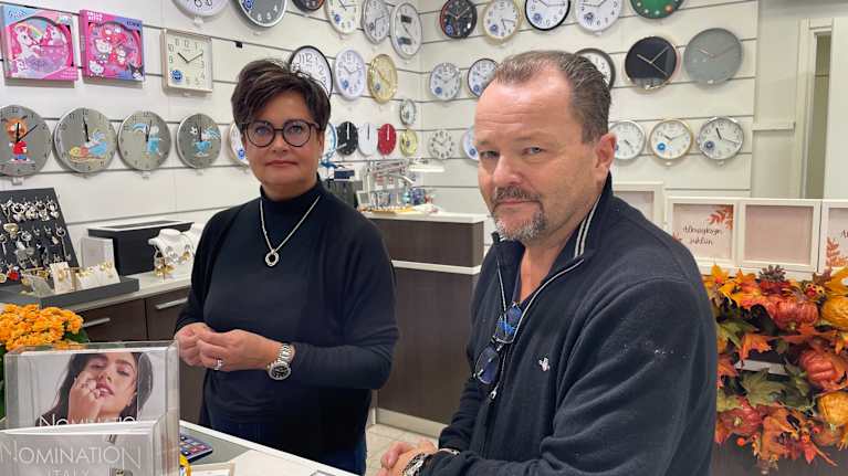 Kuvassa on kaksi henkilöä kultakaupan tiskillä, taustalla näkyy monia eri tyylisiä kelloja seinällä. 