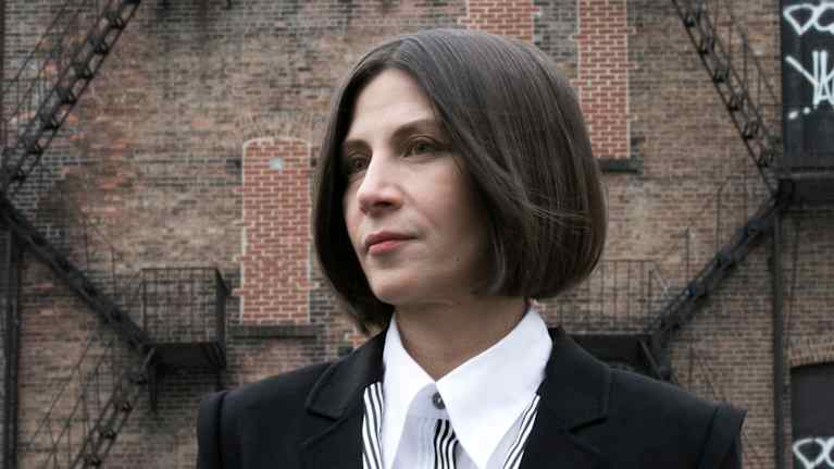 Polkkatukkainen nainen seisoo kameran edessä ja katsoo vasemmalle kaukaisuuteen.