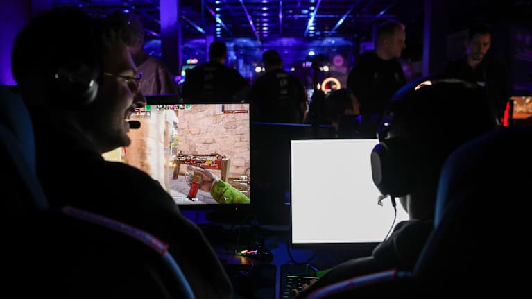 Tietokonepelin pelaajia pelitapahtumassa. Ruudulla näkyy pelihahmon käsi ja ase. Taustalla näkyy sinisiä valoja.