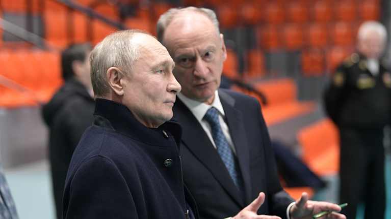 Vladimiri Putin kuuntelee Nikolai Patruševia hallissa. Putinilla on tumma päällystakki, Patrušev on mustassa puvussa ja sinisessä kravatissa.