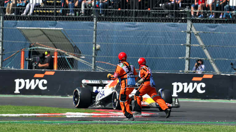 Kaksi järjestäjää juoksuu radalla, kun F1-auto lähestyy mutkaa.