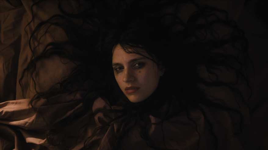 Mustahiuksinen valkoinen nainen makaa hiukset levinneinä kasvojensa ympärille, katsoo kameraan.