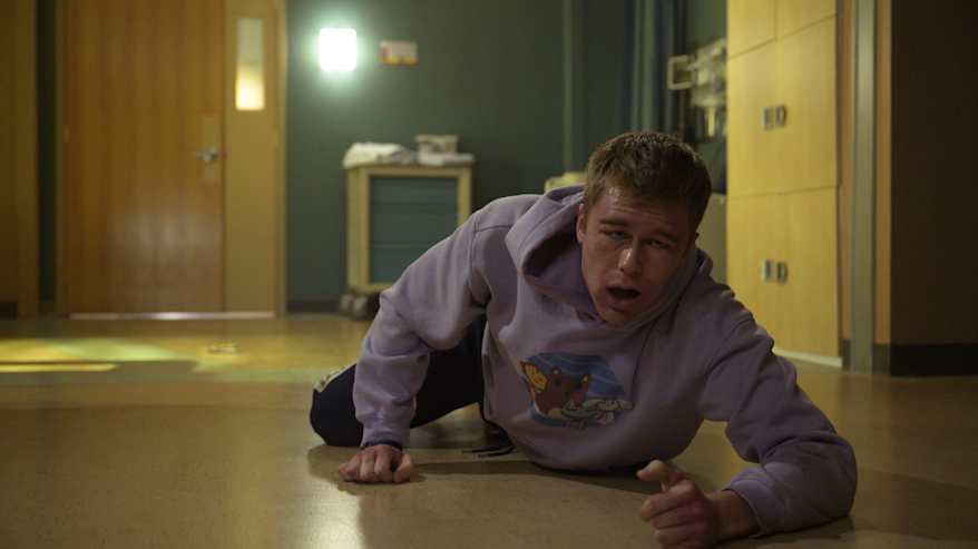 Lilaan huppariin pukeutunut nuori mies raahautuu lattialla sairaalassa.