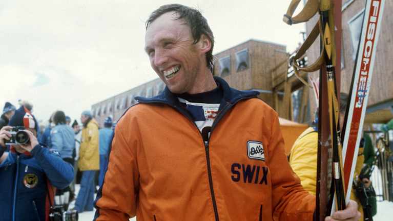 oranssitakkinen mies hymyilee sukset kädessä.