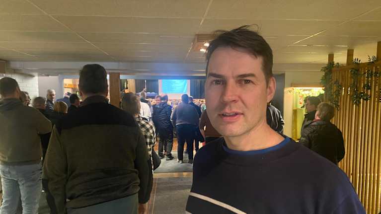 Mies seisoo rakennuksen aulassa. Hänen takanaan on iso joukko ihmisiä, joista osa seisoo, osa istuu.