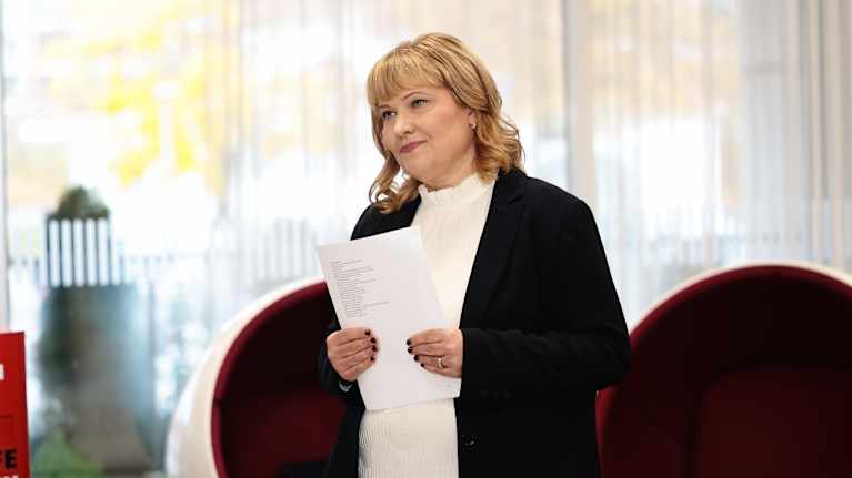 Nainen pitelee paperiarkkia käsissään ja seisoo punaisen nojatuolin edessä.