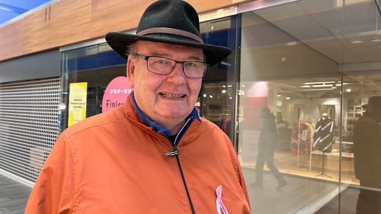 Mies seisoo kauppakeskuksen käytävällä oranssi takki ja lierihattu yllään.