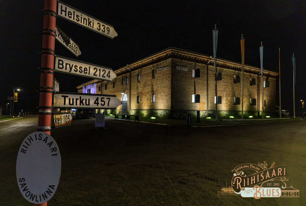 Vanha tiilinen museorakennus hämärässä illassa. Valot valaisevat tiiliseiniä. Edessä vasemmalla vanha tieviitta, jossa kaupunkien kuvia.