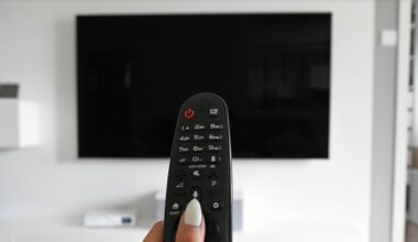 Taustalla iso musta televisioruutu ja etualalla kaukosäädin, jota sormet painavat