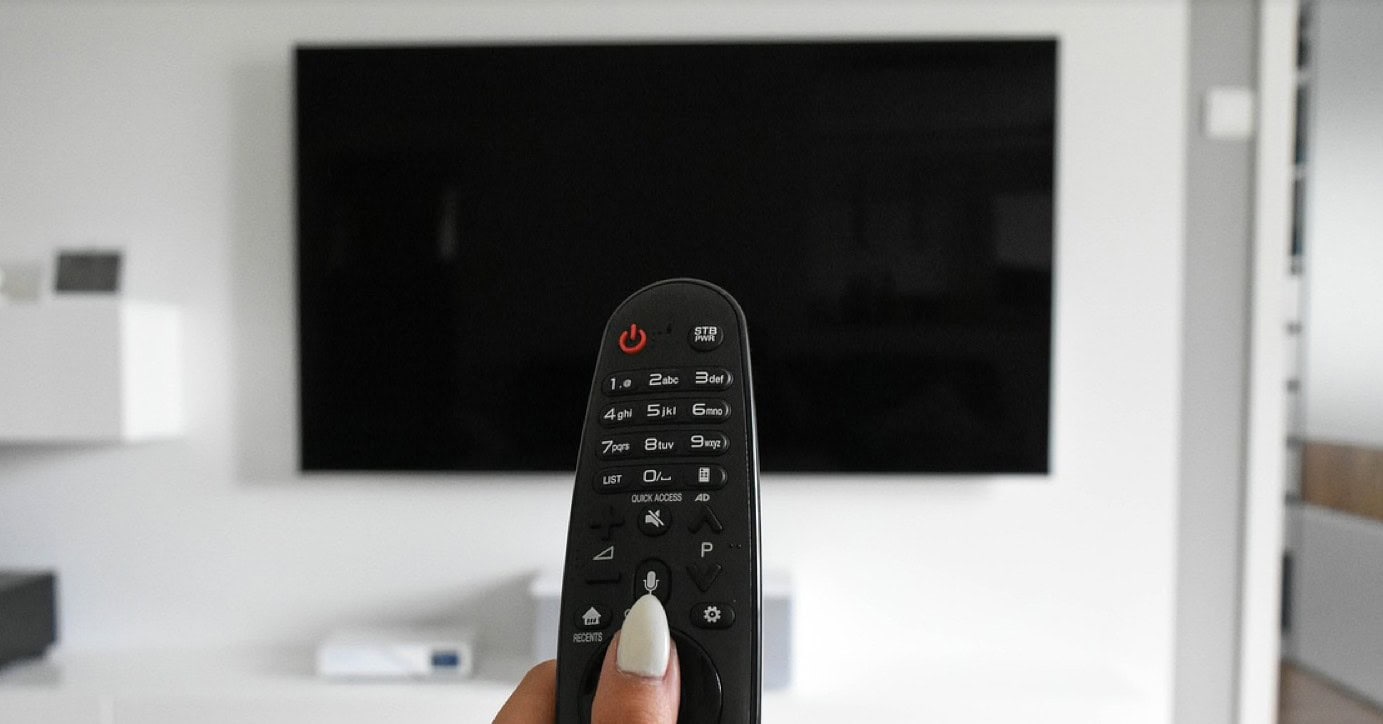 Taustalla iso musta televisioruutu ja etualalla kaukosäädin, jota sormet painavat