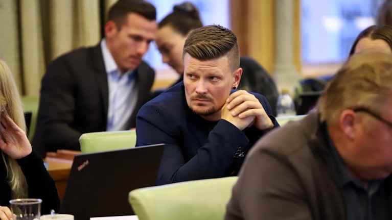 Mies istuu kädet yhdessä kasvojen edessä. Miehellä on viikset ja parta. Hän katsoo sivuun. Taustalla kaksi ihmistä keskustelee.